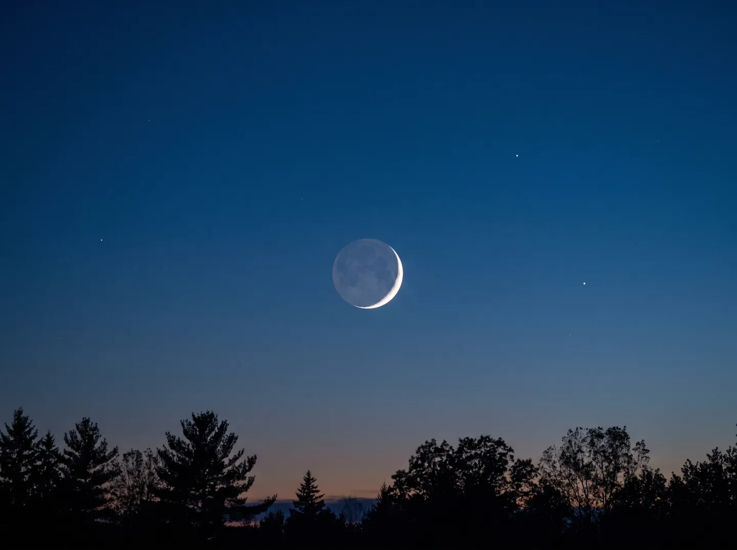 A thin crescent moon in a deep blue twilight sky, its dark part glowing faintly
